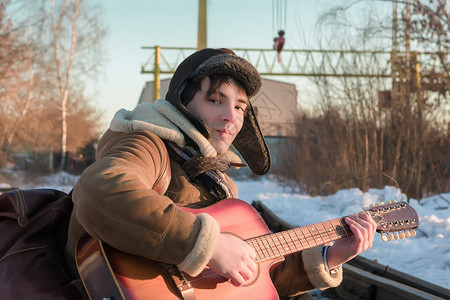 身穿羊皮大衣和戴耳巾帽的青年男子冬季在火车轨上弹吉他的冬季游戏白种人客情绪图片