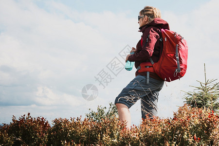 读穿着背包在山上徒步的妇女在远离自然的地方度暑假站在山顶欣赏地风景的女人拿着瓶装水的玻璃杯全景踪迹图片