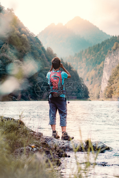 站立带着背包的年轻旅游者在山峰上看望远镜站在河边的岩石上男孩在山度假与背包一起游荡他穿着运动夏季服装笑声波兰男人图片