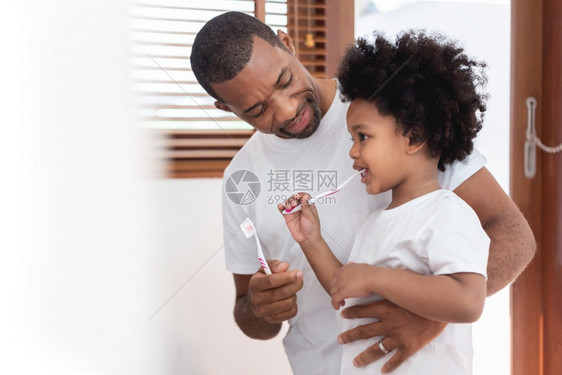 男人教学牙齿快乐的非裔美国人家庭爸教他的儿子穿着白衬衫在浴室刷牙一起在浴室里刷牙爸和孩子男卷发早上在家享受牙科卫生口腔图片