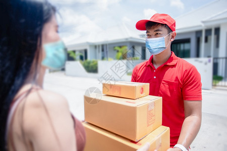 亚洲送货服务人员身穿红色制服戴帽子和面罩的亚洲送货服务人员在家中门前向女顾客提供纸板箱面罩上装着红色帽子和纸箱在线购物和快递大流图片