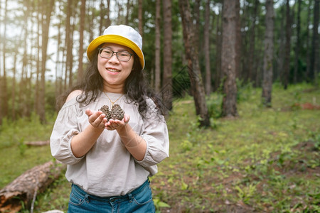 夏天针叶保持身着白礼戴帽子的亚洲女游客在自然小径森林公园上持松树绿中的锥图片