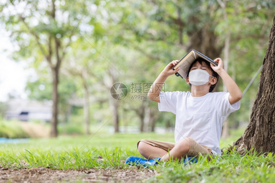 一名戴着卫生面具的白人男孩独自坐在一棵树下看着本书蒙住他的头部污染学校年级图片