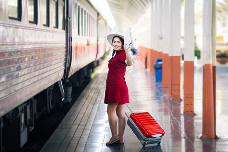 在火车站旅行中身着红裙子的亚裔孕妇持有并举手与红色行李箱打招呼女怀孕保持图片
