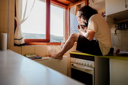 生活随意的赤脚年轻男子通过窗户用智能手机拍照同时在家中分娩吃早餐因为有冠状动脉年轻男子通过家里的窗户拍照青年男子通过他家的窗户拍图片