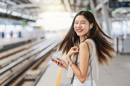年轻亚洲女乘客通过智能手机监听音乐在地铁火车站日本朝韩生活方式通勤和运输概念中采取幸福行动利用智能移电话收听音乐检查轨娱图片