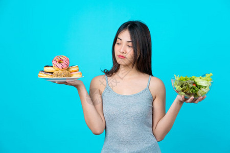 食物有吸引力的亚洲年轻女以孤立的蓝色背景体重下降和避免为饮食健康而吃垃圾食品的方式持有甜圈盘或蔬菜沙拉在玻璃碗中选择颜色协议图片