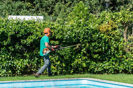 在职的一名配备刷子切割机的男在前院工作具有游泳池维护概念a园艺维护甲板正式的图片