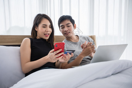冲浪坐着在家用笔记本电脑和手机支付信用卡家庭上网购物的亚洲情侣在床上冲网时快乐的一对夫妇电话图片