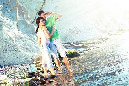 拥抱在岩石海岸的年轻夫妇站在脚踝深的水中望着地平线快乐的多岩石图片