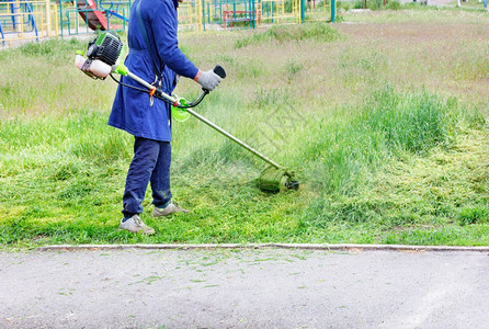 蓝衣工人在沥青道路附近的一个城市公园里用工业汽油修剪机铲草坪复制空间一个穿蓝色大衣的工人在绿色草地上磨布沿着沥青道路用汽油修剪机图片