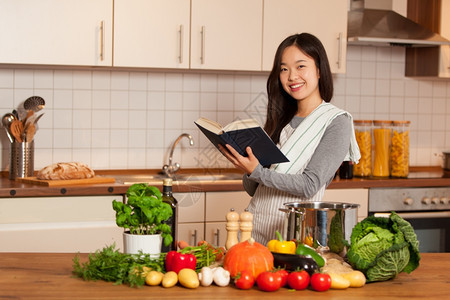 食物站在厨房的亚洲微笑着女人在看烹饪书的时候读有机图片