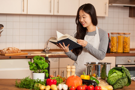 胡椒屋番茄站在厨房的亚洲微笑着女人在看烹饪书的时候图片