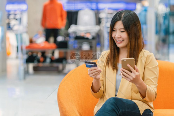 沙发小样亚洲妇女利用信卡和移动电话在百货商店通过服装背景技术钱财包和网上支付概念信用卡模型在线购物亚洲妇女使用信卡和移动电话在服图片