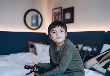 女士年轻的屋坐在床上快乐男孩拿着遥控器坐在床上CuteKitt带着微笑的脸看外面孩子在家里放松卧室看电视图片
