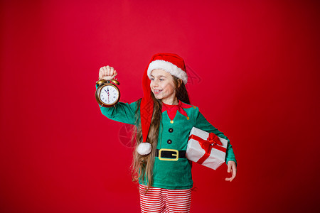 圣诞快乐迷人的女孩带着时钟和礼物穿着鲜红色亮背景的圣诞老人助手服装一个美丽的精灵婴儿肖像复制空间亮红色背景上的帮手游戏微笑魔法图片