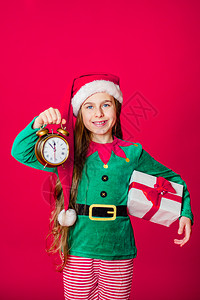 快活圣诞节乐迷人的女孩带着时钟和礼物穿着鲜红色亮背景的圣诞老人助手服装一个美丽的精灵婴儿肖像复制空间亮红色背景上的帮手帽子图片