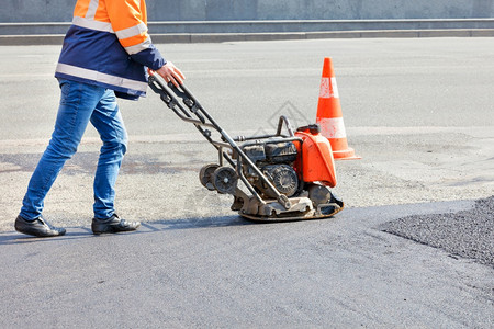 引擎地点道路工人用汽油振动板压实机在车道上沥青路工人在维修过程中用汽油振动板压实机沥青栅栏图片