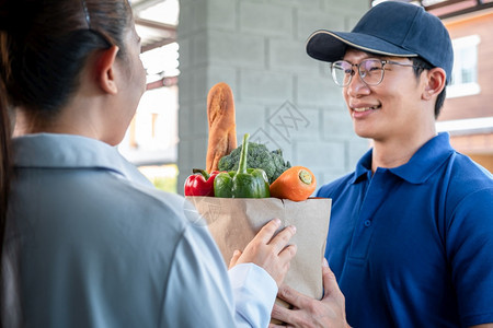 接受者妇女顾客站在家里接受一个食品水果蔬菜杂货袋由穿蓝制服的亚洲人送货员提供在科罗纳Coronavirus19流行期间提供服务带图片