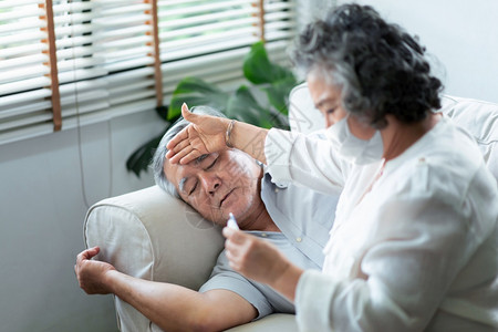 生活成熟亚洲老年病人在睡椅上躺着流感而他的妻子面戴具在温度计Corona或Covid19大流行病疾老年人寒冷时保持和检查温度寒冷图片
