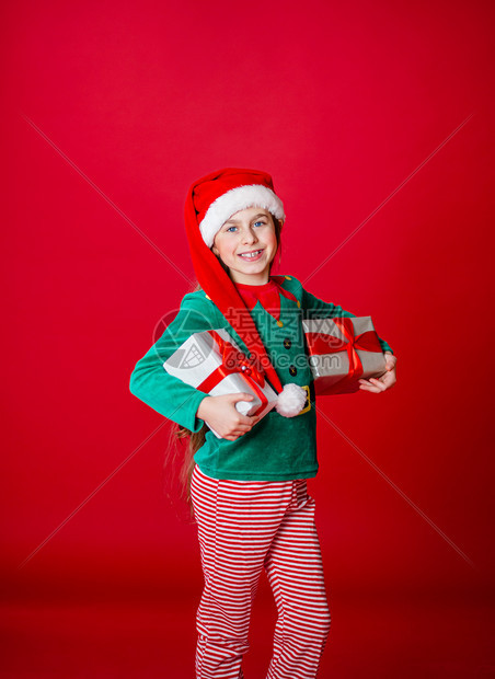 红色的圣诞快乐迷人的女孩带着礼物穿鲜艳的红色亮背景上的圣诞老人助手精灵服装一个美丽的精灵婴儿肖像复制空间圣诞快乐带着礼物的快乐迷图片