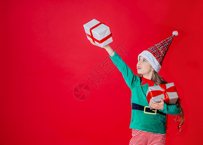 圣诞快乐迷人的女孩带着礼物穿鲜艳的红色亮背景上的圣诞老人助手精灵服装一个美丽的精灵婴儿肖像复制空间圣诞快乐带着礼物的快乐迷人女孩图片