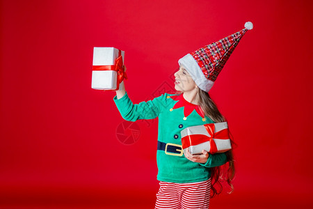 愉快白种人圣诞乐迷人的女孩带着礼物穿鲜艳的红色亮背景上的圣诞老人助手精灵服装一个美丽的精灵婴儿肖像复制空间圣诞快乐带着礼物的快乐图片