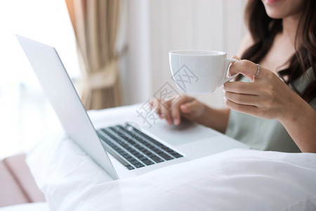 互联网年轻的在卧室枕头上使用电脑笔记本的枕头上用手提电脑时持有咖啡杯的临时妇女成年青在家工作或早上线购物自由游轮生活方式和技术概图片