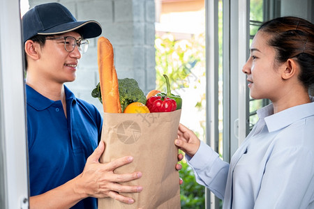 杂货店女士交身穿蓝色制服的亚洲送货男子在Corona共19流行期间服务的房子前面向女顾客提供食品水果和蔬菜杂货袋图片