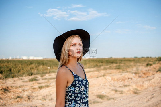 打印一位年轻美丽的caucasian金发女孩的肖像她穿着蓝色裙子印花粉和黑帽子站在沙乡村公路上夏天日落时靠近葡萄田一种苗条的图片