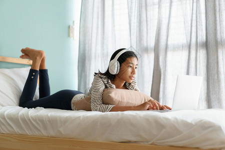 学生技术早晨亚洲女孩在清和阳光下躺在家中床上时使用带耳机的笔记本电脑快乐的亚洲女面部微笑图片
