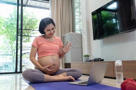 正在做偏僻的请享用美丽亚洲孕妇在家中怀时笔记本电脑上进行视频话快乐笑的年轻母亲在做瑜伽锻炼时享受聊天和与家人握手图片