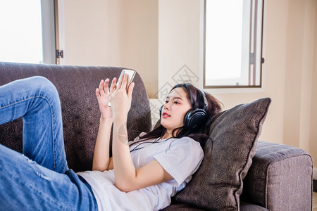 亚洲年轻快乐可爱美丽的女孩佩戴耳机听手里的音乐她正在打智能手机躺在沙发上家中睡着长椅闲暇说谎图片