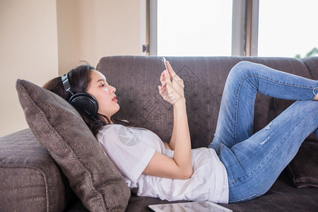 技术亚洲年轻快乐可爱美丽的女孩佩戴耳机听手里的音乐她正在打智能手机躺在沙发上家中睡着女在线的图片