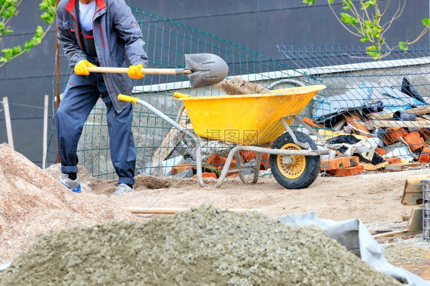 城市建造户外在施工地的背景下一名身穿深蓝内衣用铲子搬运沙的工人在一辆黄色建筑手推车中背着一把铲子装载沙的黄色建筑手推车工人用铲子图片