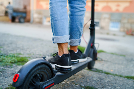 电动滑板车穿着牛仔裤和运动鞋的电脚踏车在夏日后视线上站立在人行道穿着牛仔裤和运动鞋电的输图片