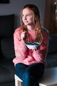 金发笑的年轻金发女孩喜欢吃新鲜蓝莓拿着一碗小水果青少年女孩穿粉红色毛衣牛仔裤坐在家里桌子上蓝色的有机女士图片