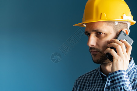 建设者在蓝墙前穿着黄色保护安全头盔和衬衫的年轻男子工程师近距离肖像隔着智能手机在电话交谈边视线上进行职业沟通图片