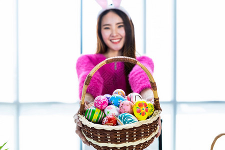 美丽的幸福复活节概念穿着兔子耳朵的亚洲快乐女青年士戴着兔子耳朵的抽象模糊与焦点节目集中展示拿着一篮子和多彩的复活节鸡蛋在白衣室背图片