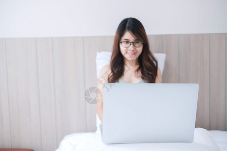 手臂坐着快乐的亚洲女坐在白床上并使用电脑笔记本年轻成女在家工作或网上购物时微笑和欢乐的年轻成女自由者生活方式和技术概念成人图片