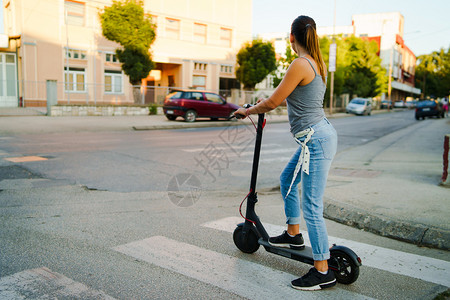 女孩在玩滑板车街道积极的年轻妇女乘电动脚踏车在街上等候夏日晚上交通中穿着牛仔裤的十字路口户外背景