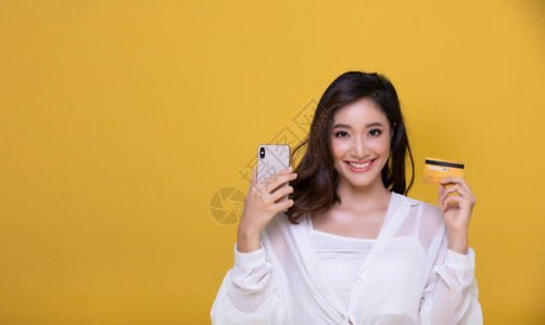 亚洲美丽的快乐年轻女微笑着欢她持有信用卡使智能电话在网上购物黄色背景幸福消费主义销售和人购物概念上买东西un手机女士移动的图片