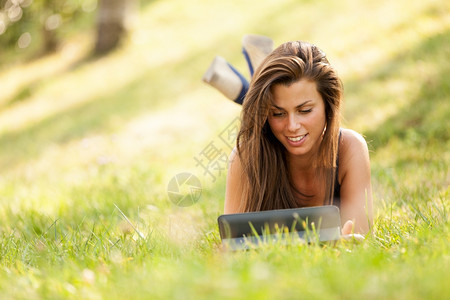 漂亮的黑褐色女孩正看着数字屏幕坐在草地上微笑着夏天药片学校图片