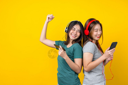 幸福派对亚洲人两个快乐的亚洲微笑年轻女她们戴着无线耳机通过智能手和孤立的黄色背景生活方式和业余爱好概念在孤立的黄色背景生活方式和图片