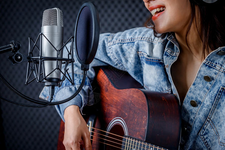一种生产快乐的笑容年轻亚洲女发声员戴着耳机吉他在专业制片室录麦克风前的歌声笑玩图片