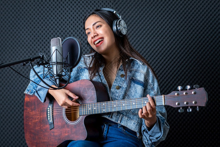 吉他手声乐专业的一位年轻亚洲女歌手戴着耳机在专业工作室录制麦克风前的歌声她笑得很高兴他开心一个年轻的亚洲女发声员戴上耳机图片