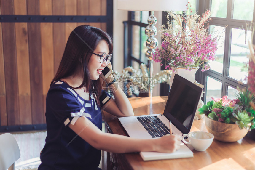 保持现代的专业亚洲女商人抓住妇女捡起智能手机作便条与笔记本电脑合作在咖啡店像背景一样在咖啡店工作图片