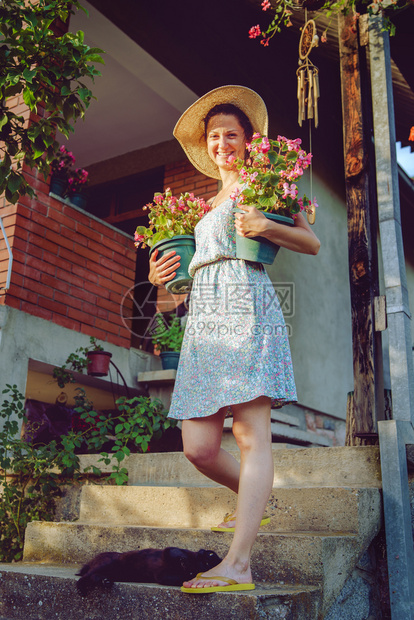 生长经过关心年轻女农民快乐笑的年轻女农民带着两壶花和个锅子在她家的花园盆旁送照顾着戴帽子和暑期服的植物图片