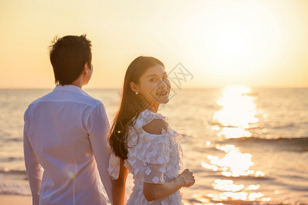 约会步行年轻的亚洲快乐情侣在海滩上握着手与一个女人面对相机鼓浪漫旅行蜜月渡暑假亚裔女和男手拥抱户外的日落背景已婚图片