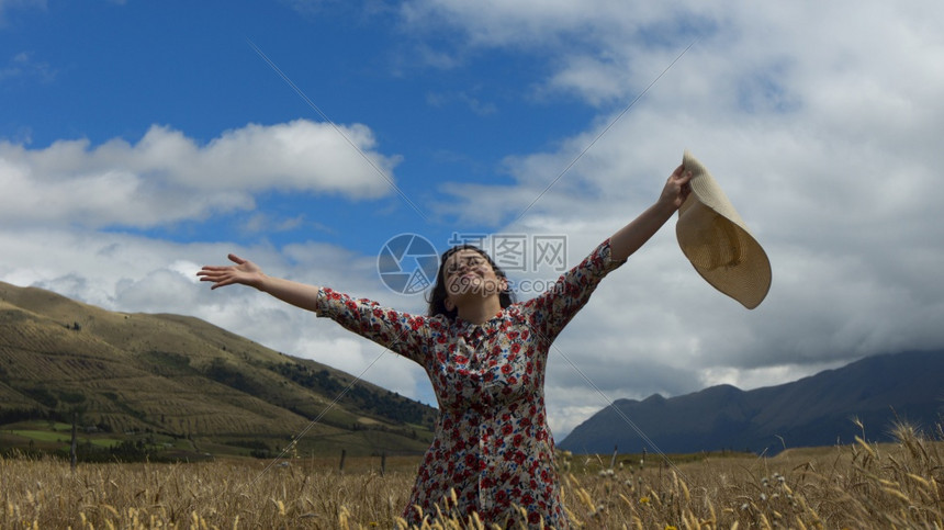 身着花朵装饰的年轻快乐女人带着高举的双臂仰望天空手拿着帽子在一片小麦田中间在阴云多的一天里蓝和山丘背景中怀着鲜花大礼服的年轻快乐图片